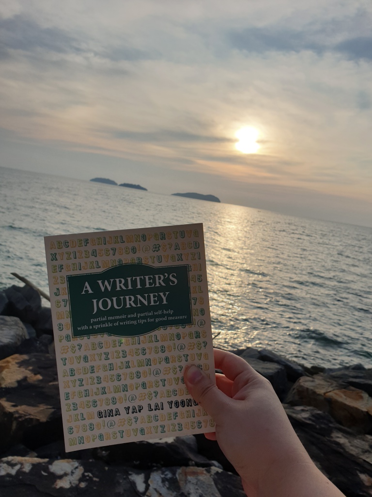 Photo of Gina’s book A Writer’s Journey with the background of the sea and setting sun.